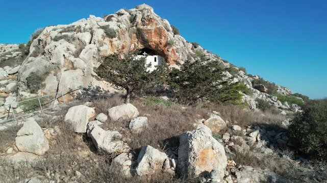 Drone scenery 4k ultra high definition of cave white Christian orthodox chapel nestled  inside a natural rock cave cavern on a rugged hillside with desert vegetation. Agioi Saranta cave church