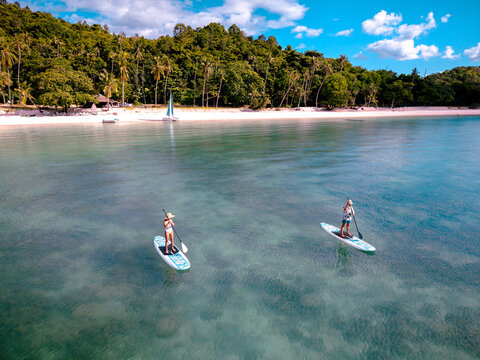 Top aerial view of couple of tourist are enjoy on water sport paddle sup-board with kayaking along at the edge of the sea beach under crystal clear of water in the island of summer trip