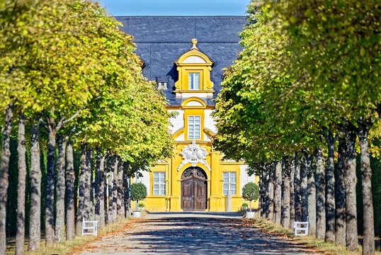 Palace garden of Seehof Castle, symmetrical avenue with chestnut trees and high hedge walls along a pebble path with autumn leaves, in the background the yellow-white fa&ccedil;ade of the baroque castle under a slightly cloudy blue sky, Memmelsdorf, Bamberg district, Upper Franconia, Bavaria, Germany