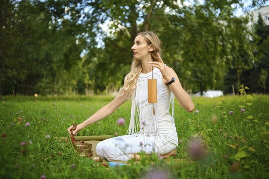 A woman sits calmly on a soft grassy area, softly striking a singing bowl while surrounded by vibrant greenery. The peaceful setting invites relaxation and mindfulness, ideal for meditation practice