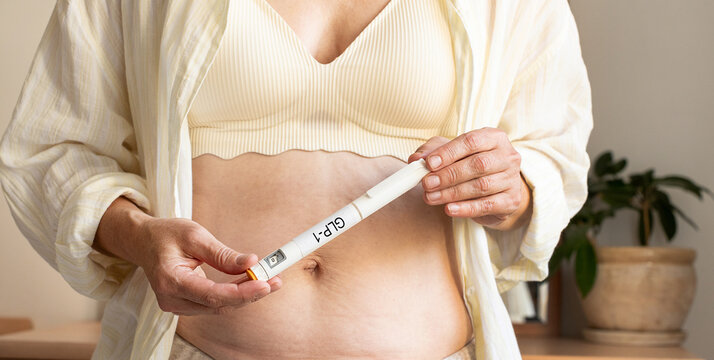 Woman holds GLP-1 pen injector against her abdomen at home. Concept of medical weight loss, obesity treatment, use of injectable medications, and body weight management