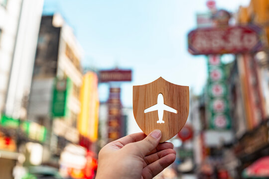 Travel insurance and flight protection concept with hand holding wooden shield with airplane cutout against bustling Asian city street market for holiday safety and overseas trip coverage.