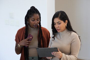 Diverse female colleagues standing together reviewing tablet while holding purple phone at office