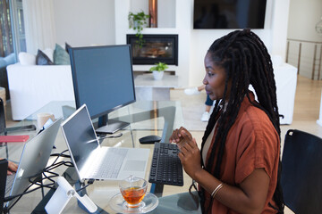 Obraz premium African American woman sitting at glass-top desk in living room using laptop and monitor