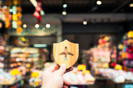 Travel insurance and tourist protection concept with hand holding wooden shield with airplane cutout against colorful souvenir shop background for holiday shopping safety and trip coverage.