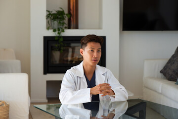 Obraz premium Mid adult Asian man sitting at living room glass coffee table wearing lab coat over scrubs