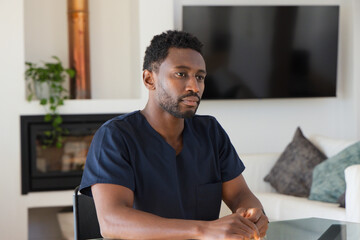 Obraz premium Mid-adult African man sitting at glass table in living room wearing navy scrubs, viewing TV