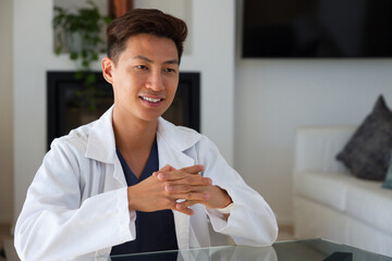 Obraz premium Mid-adult Asian man wearing white lab coat, navy scrubs sitting at glass table in living room