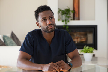 Obraz premium Mid adult African man wearing navy scrub top, sitting at table in living room, clasping hands