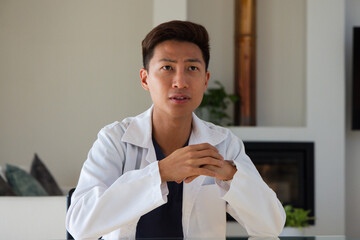 Obraz premium Mid-adult Asian man sitting at glass-topped table in modern living room wearing white lab coat