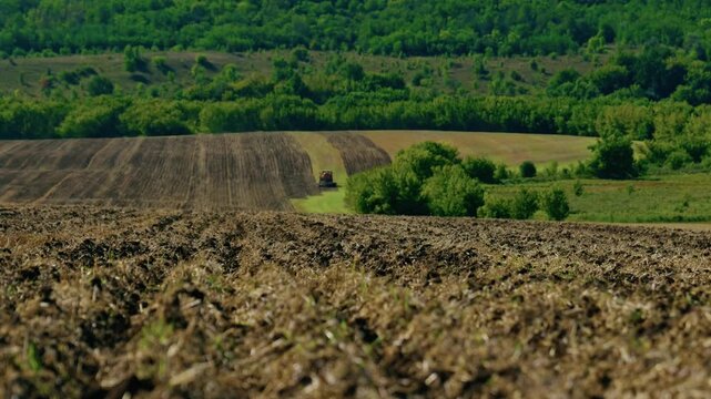 A wide landscape shot captures a plowed agricultural field with tilled furrows extending toward rolling green hills under bright sunlight.