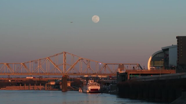 Full moon rising over river bridge and city waterfront with small plane flying across clear evening sky.