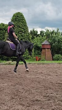 Rider practices on a black horse in a riding arena during cloudy weather