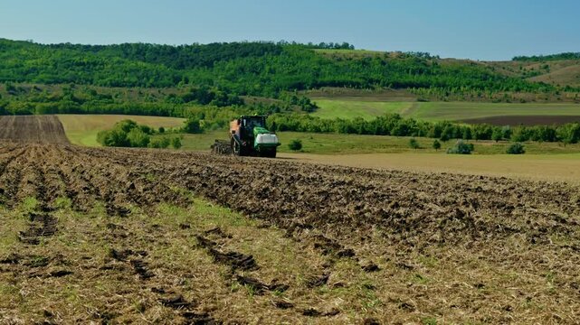A wide landscape shot of a modern tractor sowing crops across a cultivated field with scenic green hills under clear blue sky in bright daylight.