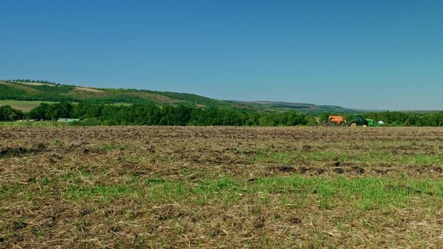 A wide landscape shot of a modern tractor sowing crops across a cultivated field with scenic green hills under clear blue sky in bright daylight.