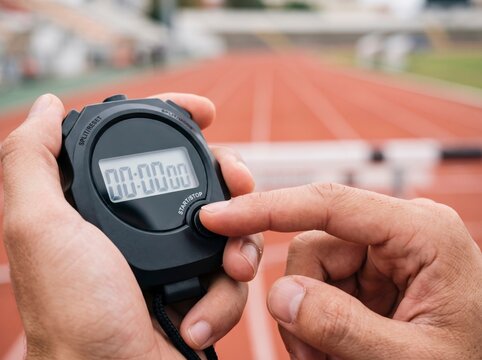 Close up of hands holding and pressing a digital stopwatch timer with a blurred red athletic running track in the background.