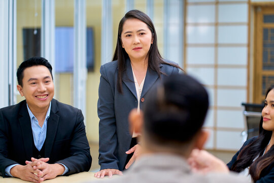 Confident Asian female executive standing and leading a corporate team meeting in a modern office. Professional Asian business woman manager listening to colleagues during board presentation.