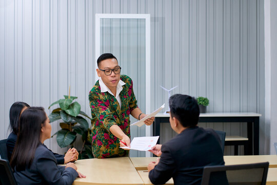 Asian businessman standing and handing financial report document to colleague during corporate team meeting in modern office. Expressive executive leading business presentation and document sharing.