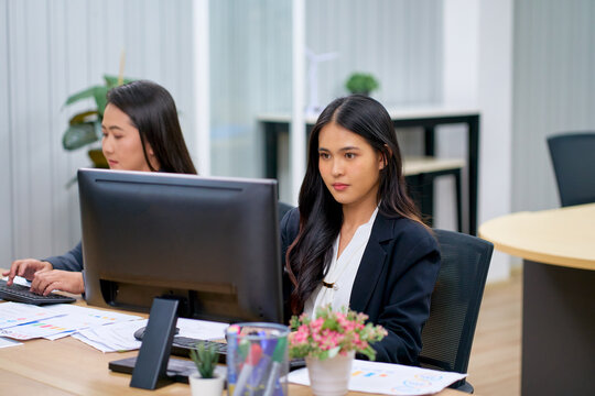 Focused young Asian businesswoman working on desktop computer at office desk. Concentrated female employee analyzing data on screen with colleague working in modern corporate workspace background.