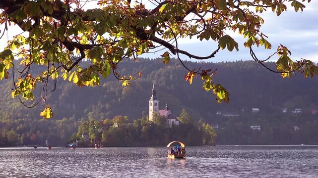 A small tourist boat slowly crosses lake bled with historic bled island church against green mountain landscape.