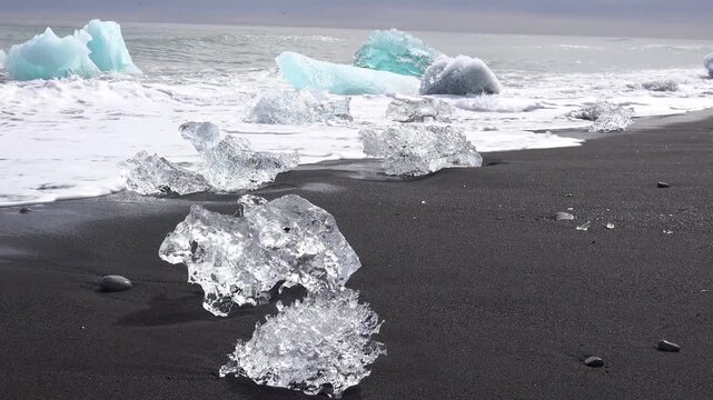 Glacier ice blocks on black sand beach with crashing ocean waves in Iceland cinematic landscape.