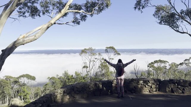 A girl hiker stands and admires the thick clouds emerging above the eucalyptus bush and a meadow, from the summit of Mount Macedon, Macedon Ranges, near Melbourne, Victoria, Australia
