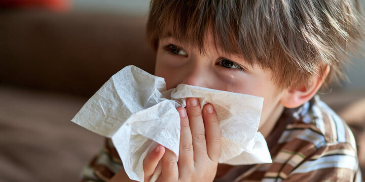 A young boy with a tissue to his nose, looking sad and unwell. He has big brown eyes and a checkered shirt