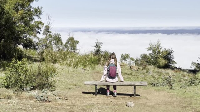 A girl hiker sits on the bench and admires the thick clouds emerging above the eucalyptus bush and a meadow, from the summit of Mount Macedon, Macedon Ranges, near Melbourne, Victoria, Australia