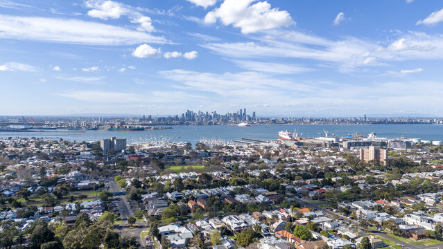 Aerial vista over the coastal suburb of Williamstown, looking across bay toward the distant city skyline, with residential house roofs, boat harbours and shipping channels. Melbourne Australia