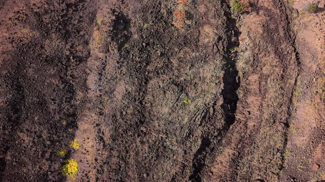 Aerial top down view of rugged volcanic lava terrain in Maui Hawaii USA with red cinder slopes dark basalt rock green shrubs and lichen patches under warm sunlight on barren crater ground.