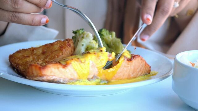 Close-up cinematic shot of woman hands slicing and serving perfectly cooked salmon steak with creamy sauce, broccoli, and hash brown on a white plate in restaurant
