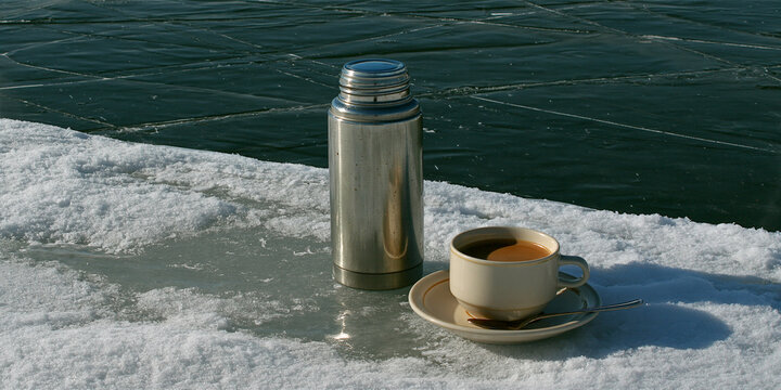 A thermos and a cup of hot coffee sit on the ice during a winter day, representing warmth and comfort in the cold