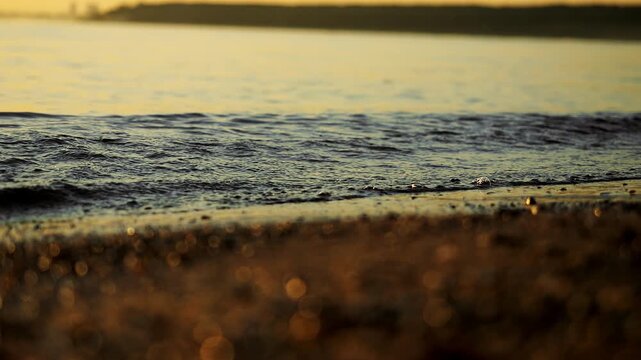 Close-up of a gentle wave rolling onto a sandy shore in warm golden sunset light. Calm water, sparkling reflections and soft natural beach atmosphere.
