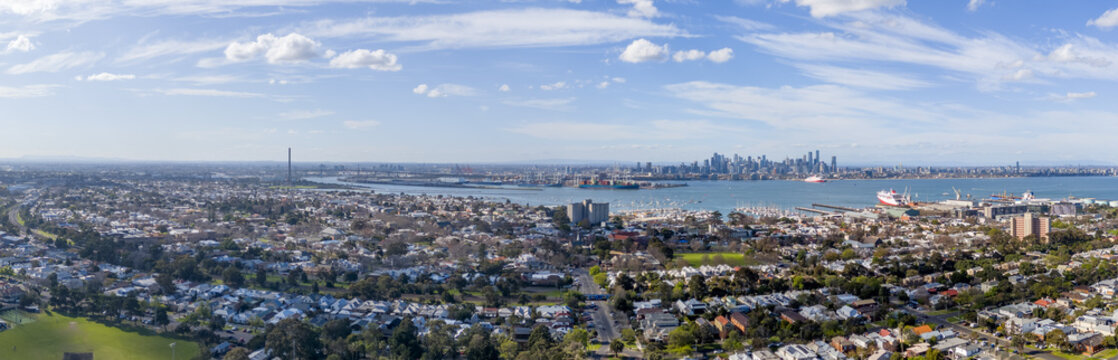 Wide panoramic aerial view over Williamstown and Newport across the Yarra River mouth toward CBD skyline, featuring residential houses, industrial stacks, cargo ships. Melbourne Australia