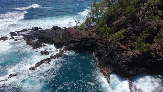 Aerial view of Kaihalulu Beach on Maui Hawaii island with white waves crashing over black lava rock cliffs, red volcanic soil and green trees above deep turquoise and blue ocean water.