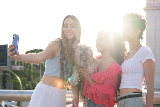 Young women friends smiling, relaxing, and taking a selfie with a small dog outdoors during sunset