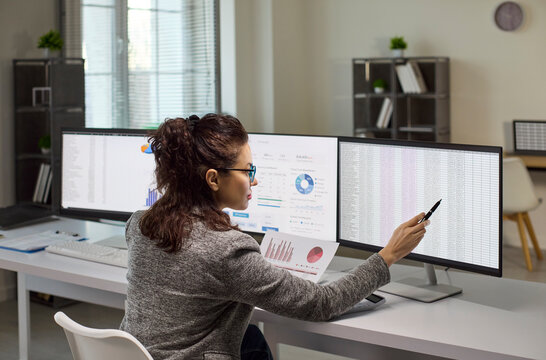 Analyst woman office computer monitors finance data spreadsheet. She reviews a report on dual screens and points at figures to perform audit. Symbolizes precise analysis and financial control.