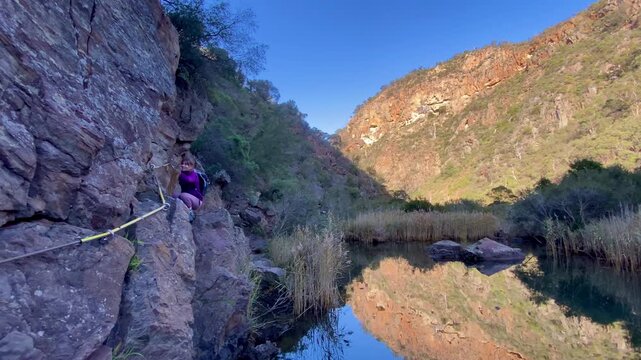 A girl hiker rock scrambles along a cliffside, holding the cable handrail above the river, on a tough section of the Werribee Gorge Circuit Walk, near Melbourne, Victoria, Australia.