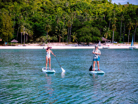 Top aerial view of couple of tourist are enjoy on water sport paddle sup-board with kayaking along at the edge of the sea beach under crystal clear of water in the island of summer trip