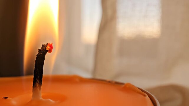 Intimate detail of a glowing flame rising from a charred wick in melted tangerine wax beside a softly lit sheer curtain, creating warm household ambience with generous neutral copy space area