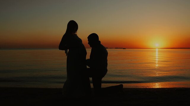 Silhouette of an expecting father kneeling beside a pregnant woman and gently touching her baby bump at sunset. Emotional maternity scene by calm water.