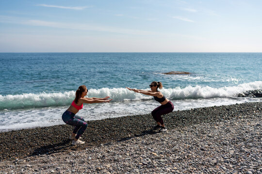 Squatting women perform outdoor fitness training on a sunny pebble beach shoreline during a coastal workout session