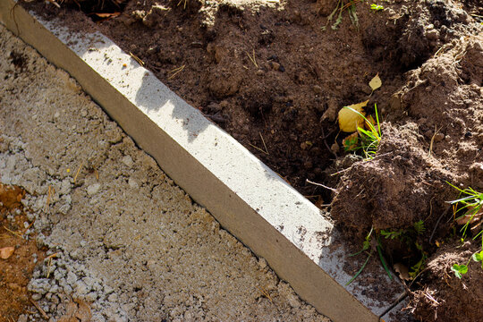 Installation of a new concrete curbstone along a garden path construction site, showing detailed stone texture and surrounding fresh soil during landscape improvement work