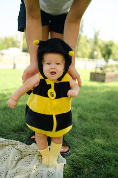 A mother holds her baby, who is dressed in a bumblebee costume, and teaches him to take his first steps on the grass in the park; a theme of childhood.