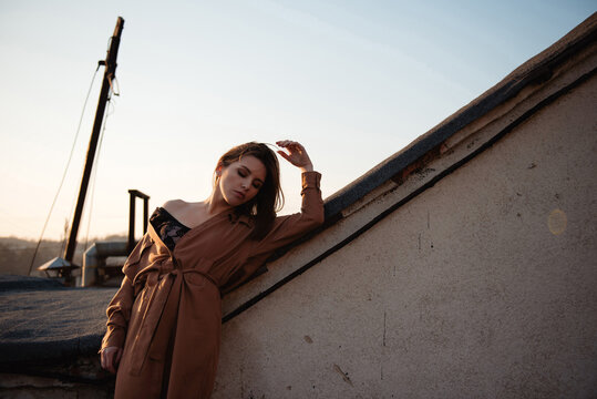 Young woman in a brown coat posing on a rooftop at sunset