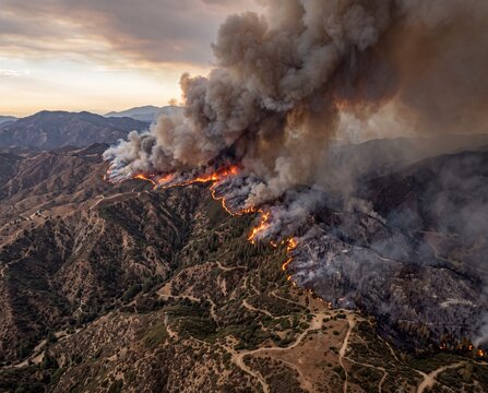 Massive wildfire raging across dry mountain slopes with thick plumes of dark smoke during sunset hours