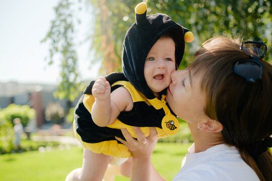 Happy mother kissing cute baby in bee costume outdoors in sunny park, loving family, motherhood and childhood concept.