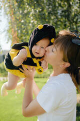 Happy mother kissing cute baby in bee costume outdoors in sunny park, loving family, motherhood and...
