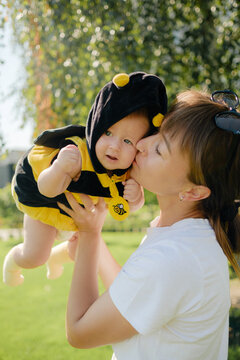 Happy mother kissing cute baby in bee costume outdoors in sunny park, loving family, motherhood and childhood concept.
