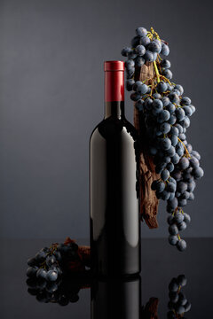 Bottle of red wine with a weathered snag and blue grapes on a black reflective background.
